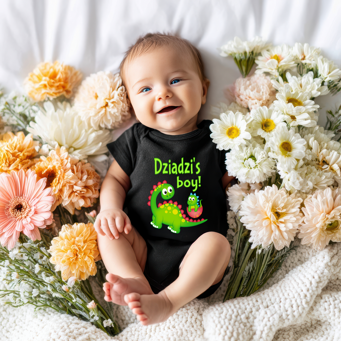A smiling baby wearing a black shirt with a dinosaur graphic lies on a bed surrounded by colorful flowers.
