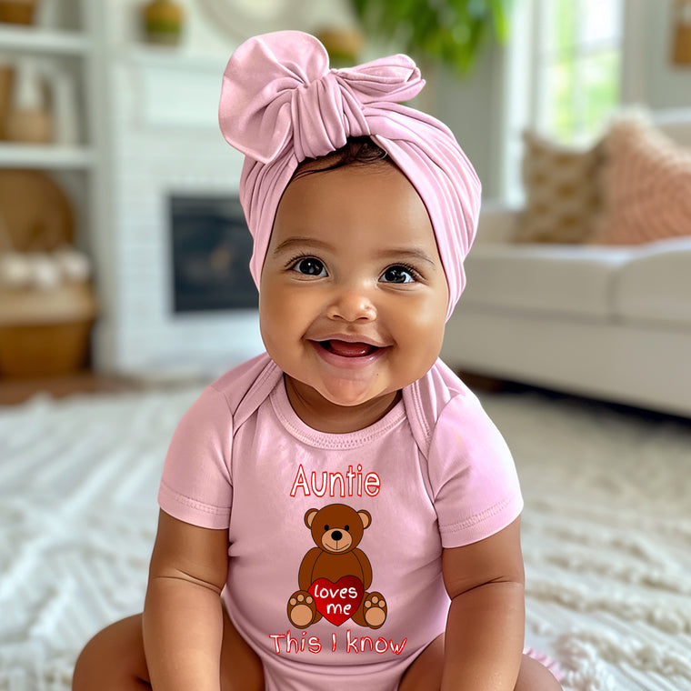 A smiling baby girl wearing a pink headband and a pink onesie with a teddy bear graphic, sitting on a white rug in a living room.