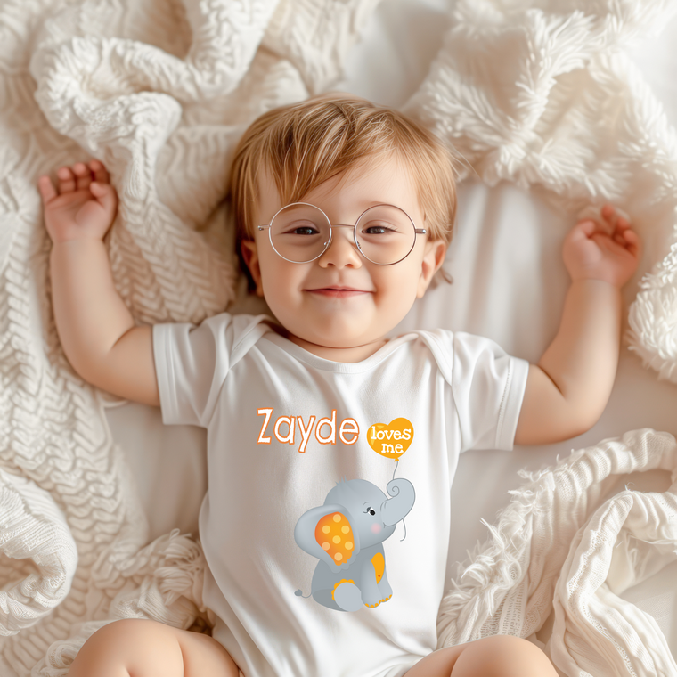 A smiling baby wearing a white onesie with an elephant design and the text "Zayde loves me" is lying on a white blanket.