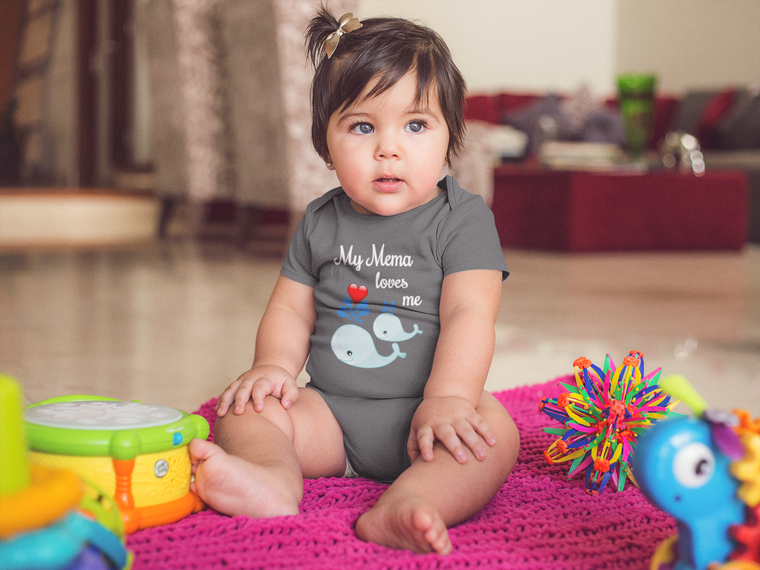 A baby girl wearing a gray t-shirt with a whale design sits on a pink blanket surrounded by toys, including a toy drum and a toy fish.