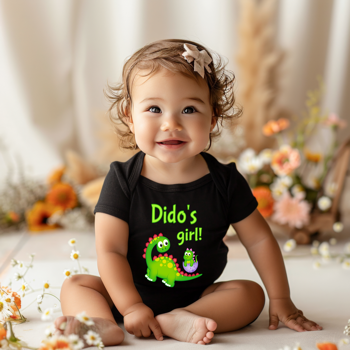 A smiling baby girl wearing a black shirt with a dinosaur graphic and the text "Dido's girl!" is sitting on the floor surrounded by flowers.