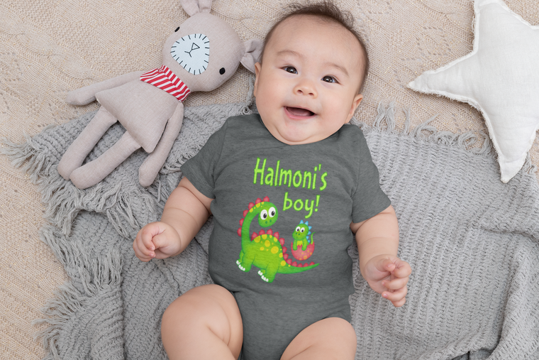 A baby wearing a grey t-shirt with a dinosaur graphic and the text "Halo's boy!" lying on a gray blanket next to a stuffed animal.
