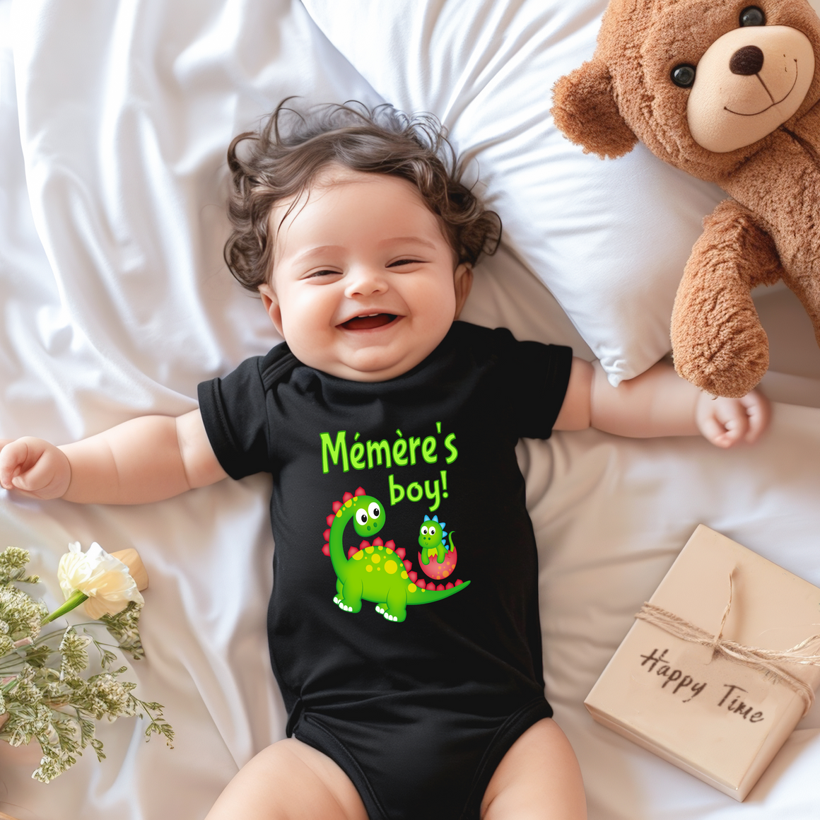 A smiling baby wearing a black t-shirt with a dinosaur graphic lies on a bed next to a stuffed teddy bear.