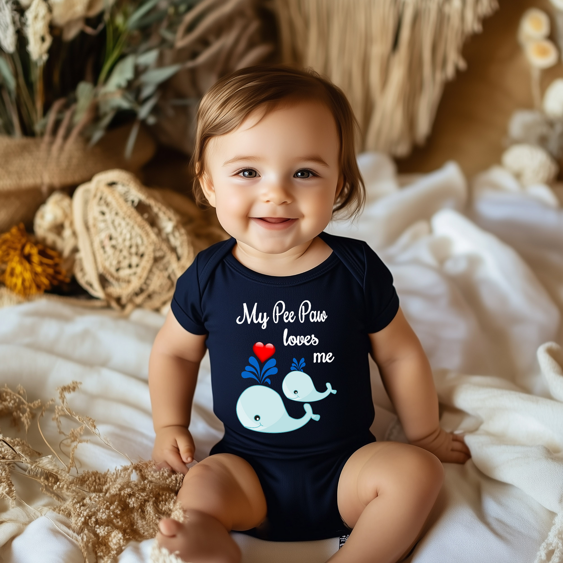 A smiling baby wearing a navy blue onesie with a whale design and the text "My Paws loves me" is sitting on a bed.