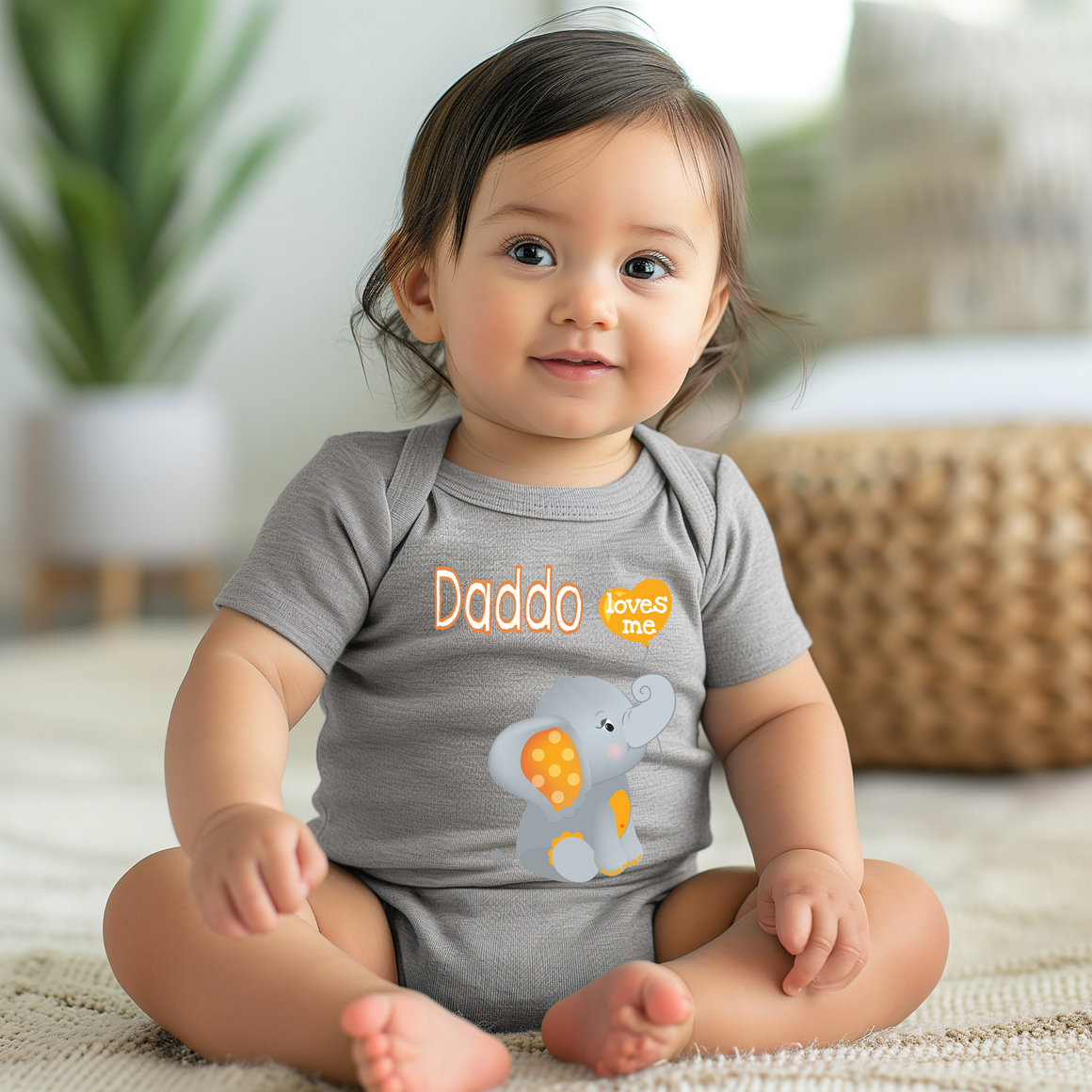 A smiling baby girl wearing a grey onesie with an elephant design sits on the floor.