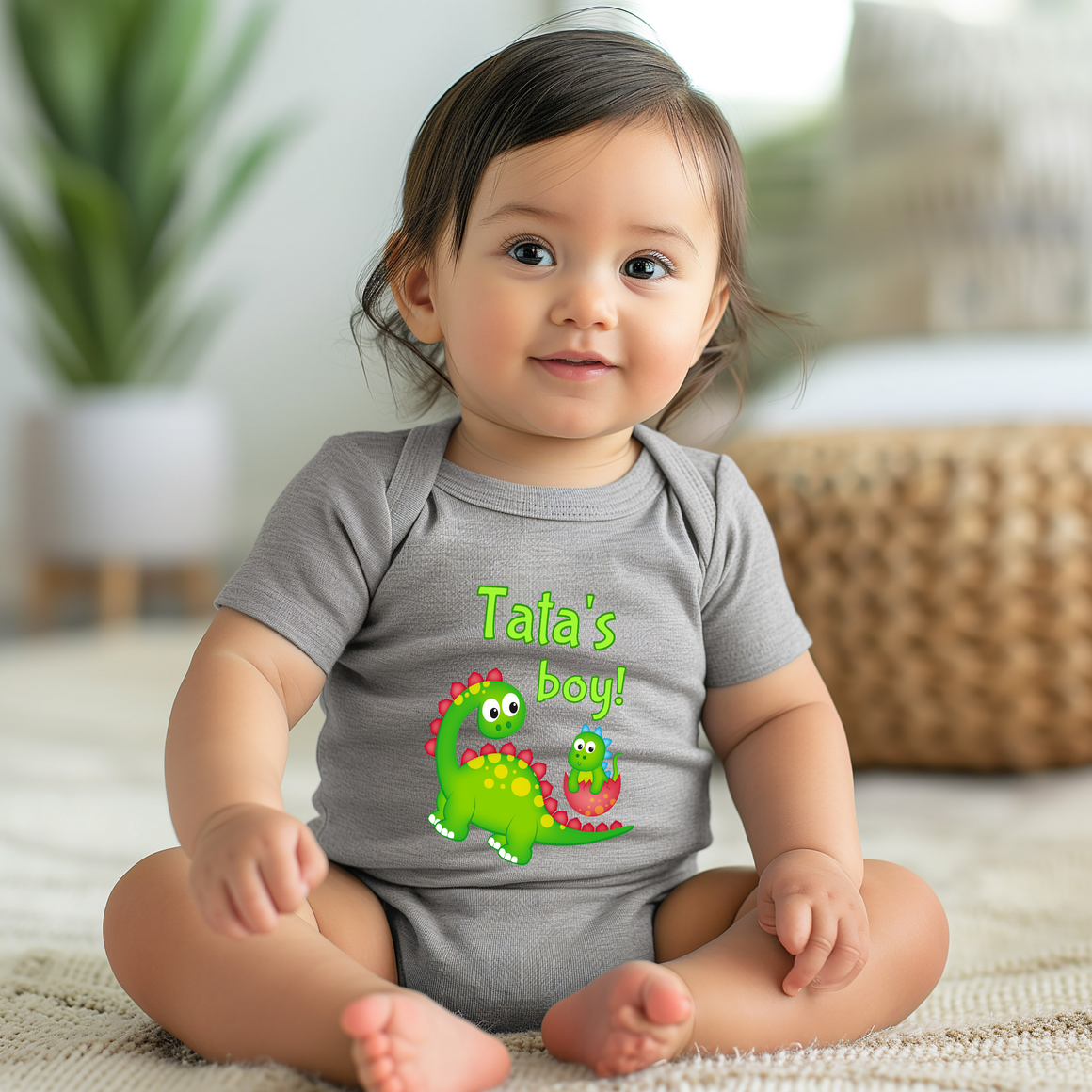 A smiling baby girl wearing a gray onesie with a cartoon dinosaur and the text "Tata's boy!"