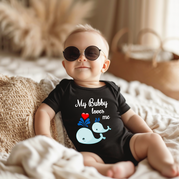 A baby wearing a black outfit with a graphic of a whale and the text "My Bubby loves me" is sitting on a bed.