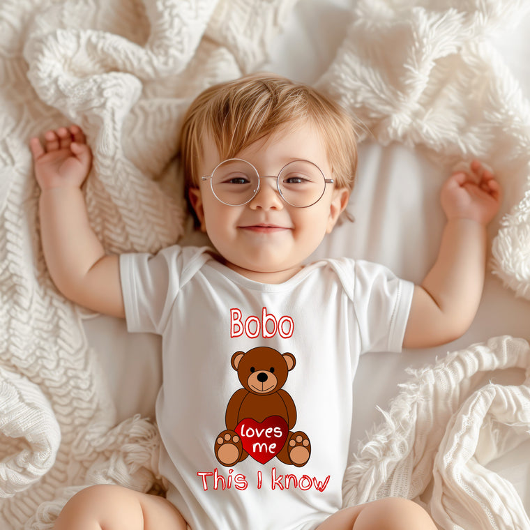 A smiling baby wearing a white onesie with a teddy bear graphic and the text "Bobo loves me This I Know" printed on it.