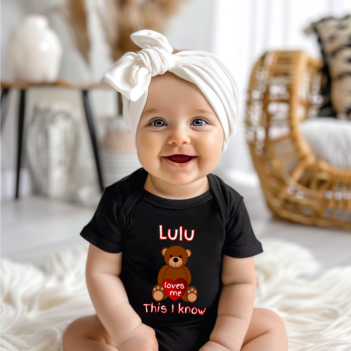 A smiling baby girl wearing a black t-shirt with a teddy bear graphic and the text "Lulu loves me This I know" is sitting on a white blanket.