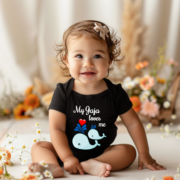 A smiling baby girl wearing a black shirt with a whale graphic and the text "My Gaya loves me" sits on the floor surrounded by flowers.