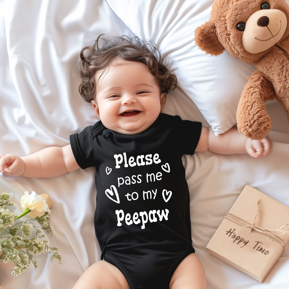 A smiling baby wearing a black onesie with the text "Please pass me to my Peepaw" lies on a bed next to a stuffed teddy bear.