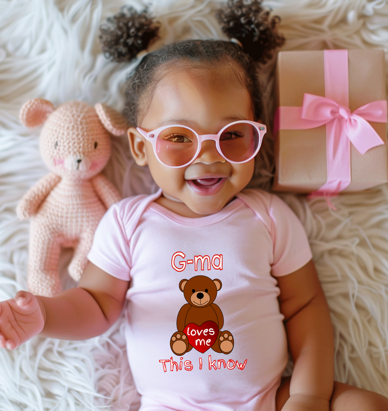 A smiling baby girl wearing a pink outfit and glasses lies on a white surface next to a pink crocheted teddy bear.