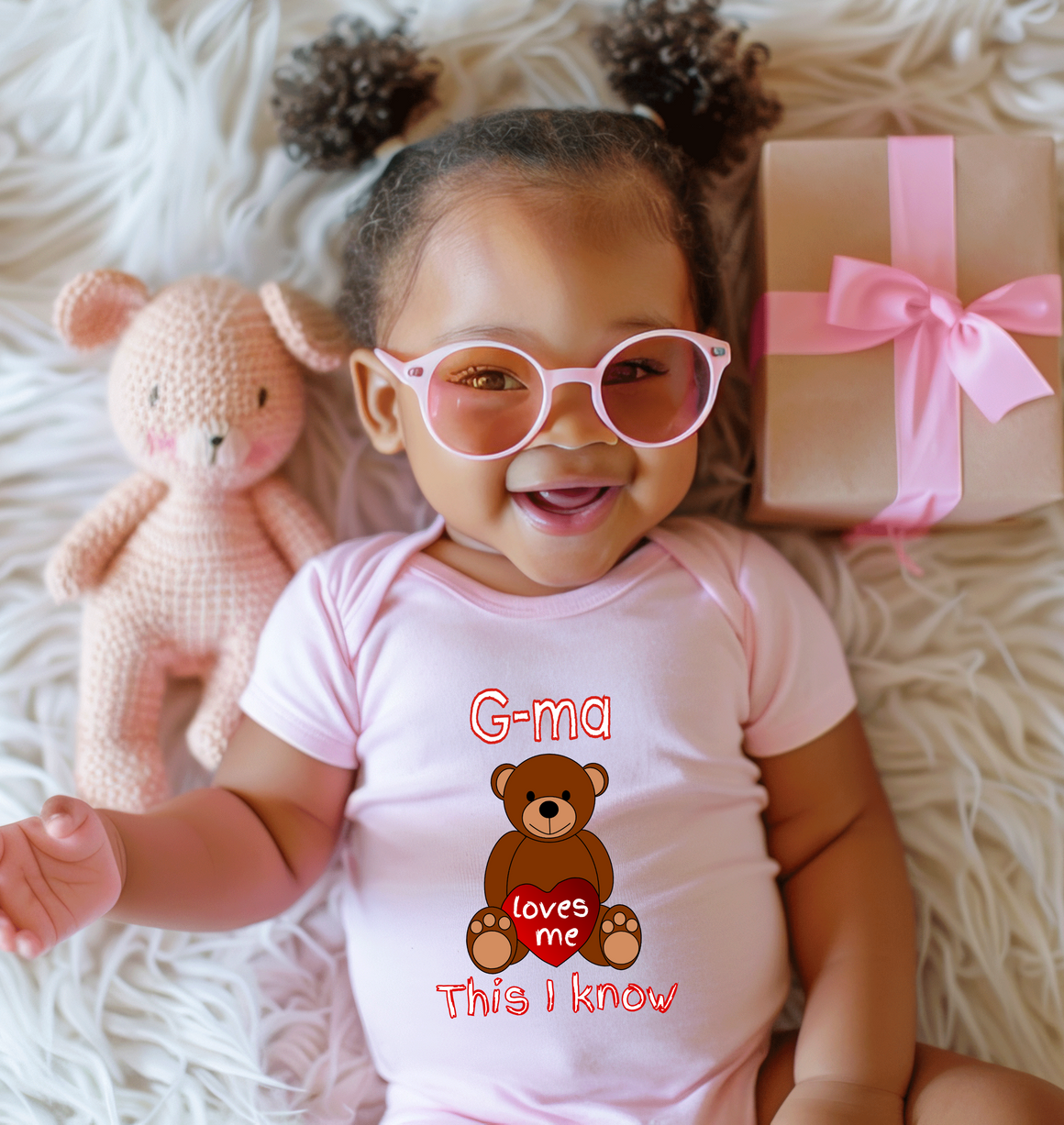A smiling baby girl wearing a pink outfit and glasses lies on a white surface next to a pink crocheted teddy bear.