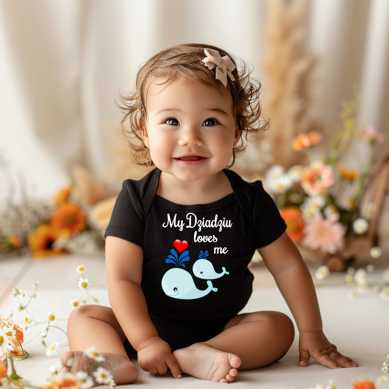 A smiling baby girl wearing a black t-shirt with a whale graphic and text, sitting on the floor surrounded by flowers.