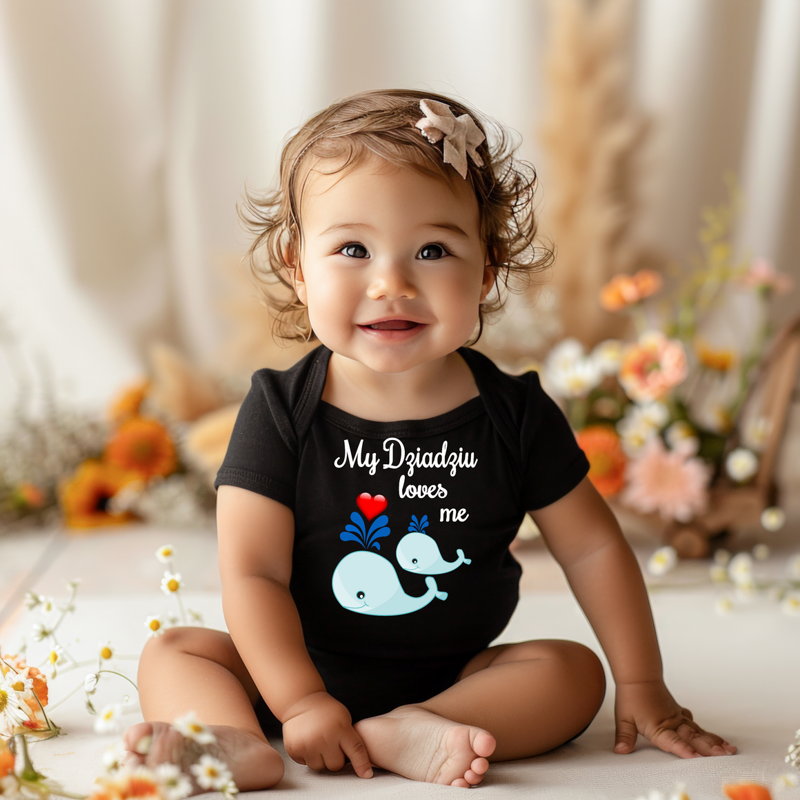 A smiling baby girl wearing a black t-shirt with a whale graphic and text, sitting on the floor surrounded by flowers.