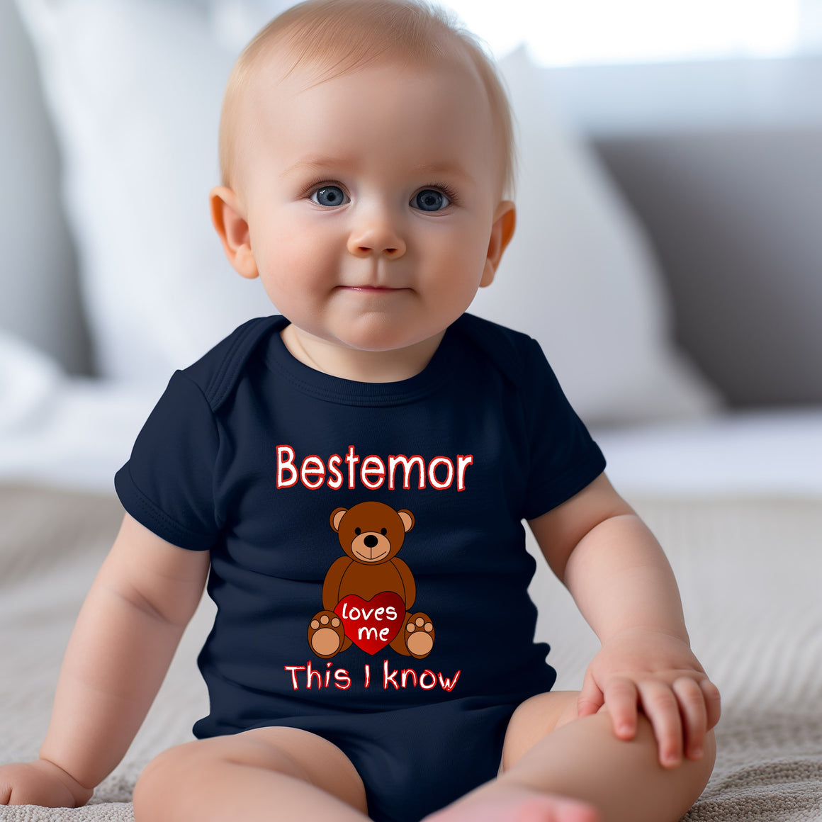 A smiling baby wearing a navy blue t-shirt with a teddy bear graphic and text that says "Bestemor loves me This I know".