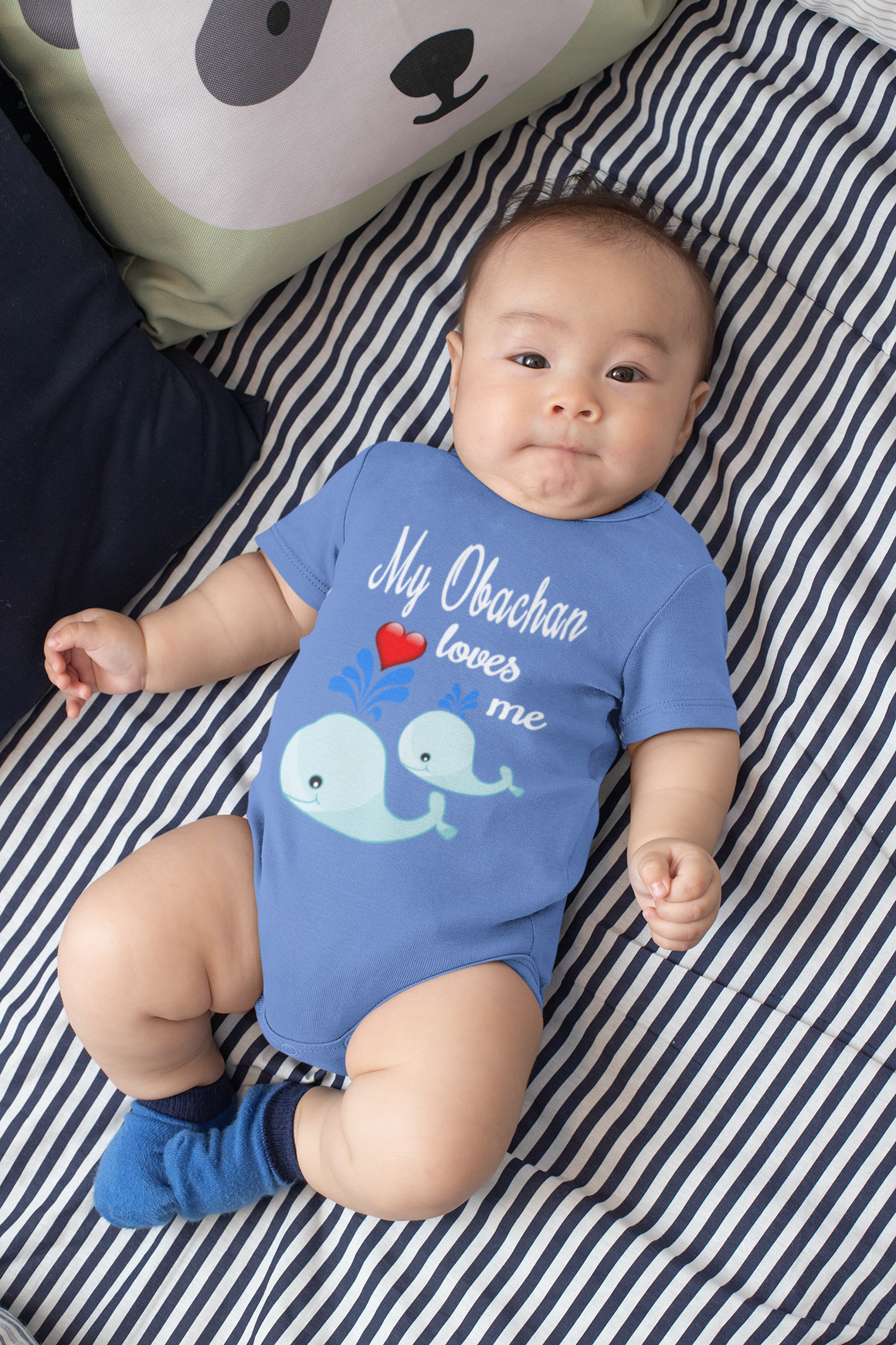 A baby wearing a blue onesie with a whale design lies on a striped bedspread.
