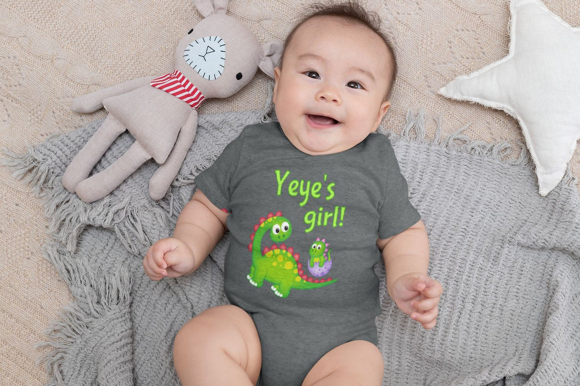 A baby wearing a gray onesie with a dinosaur and the text "Yeye's girl!" is lying on a gray blanket, smiling and surrounded by stuffed animals.