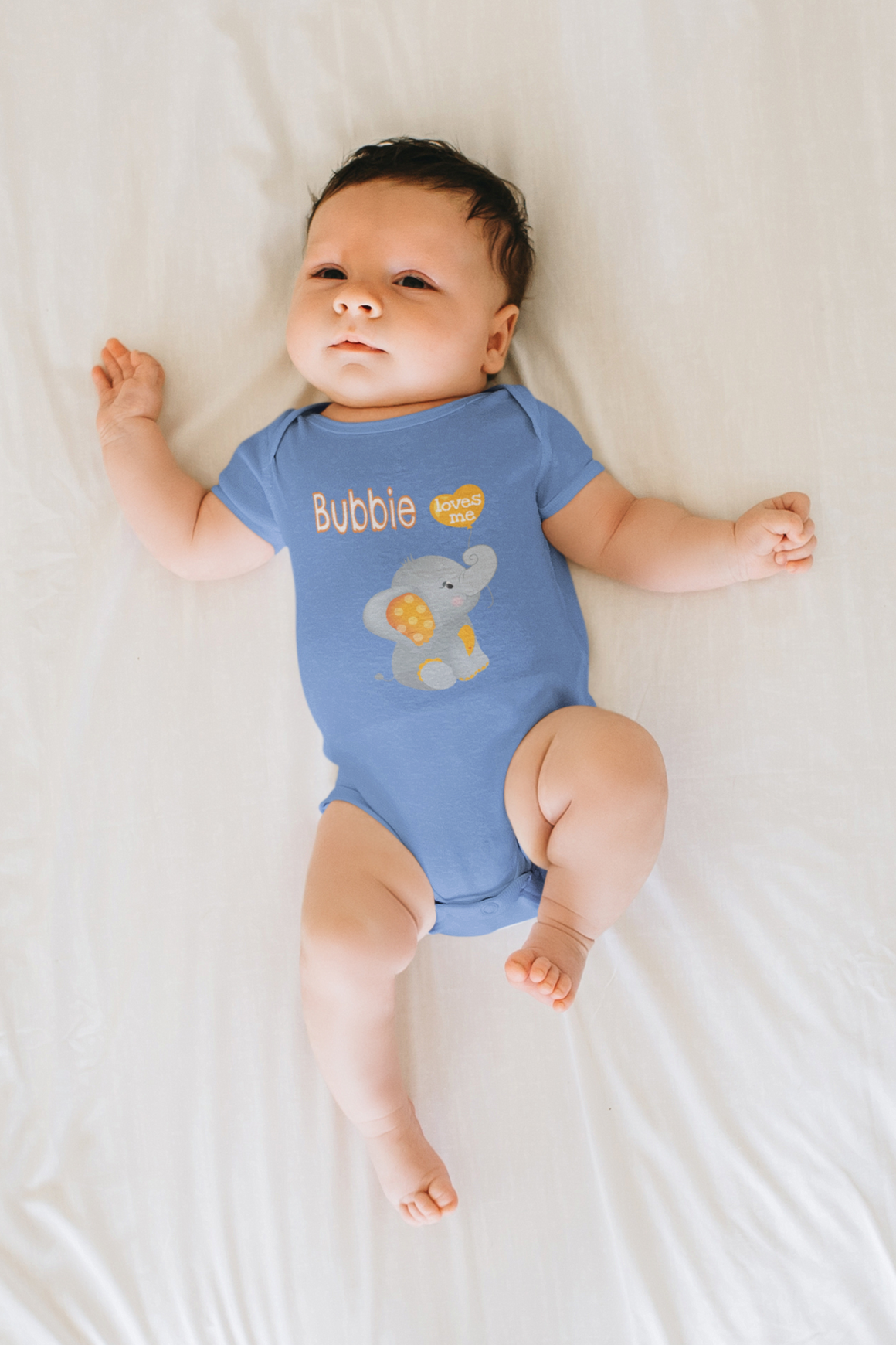 A baby wearing a blue onesie with an elephant design is lying on a white bed.