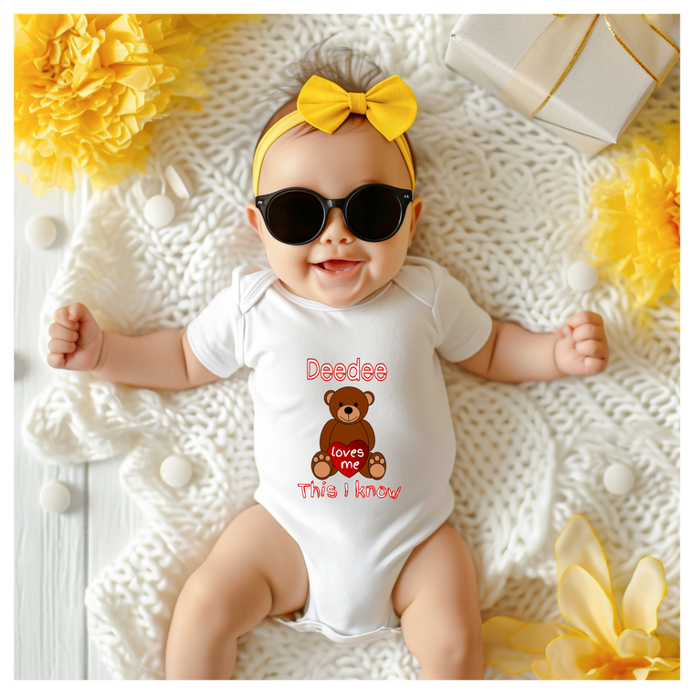 A baby wearing a white onesie with a teddy bear and the text "Deebee loves me" is lying on a white blanket surrounded by yellow flowers.