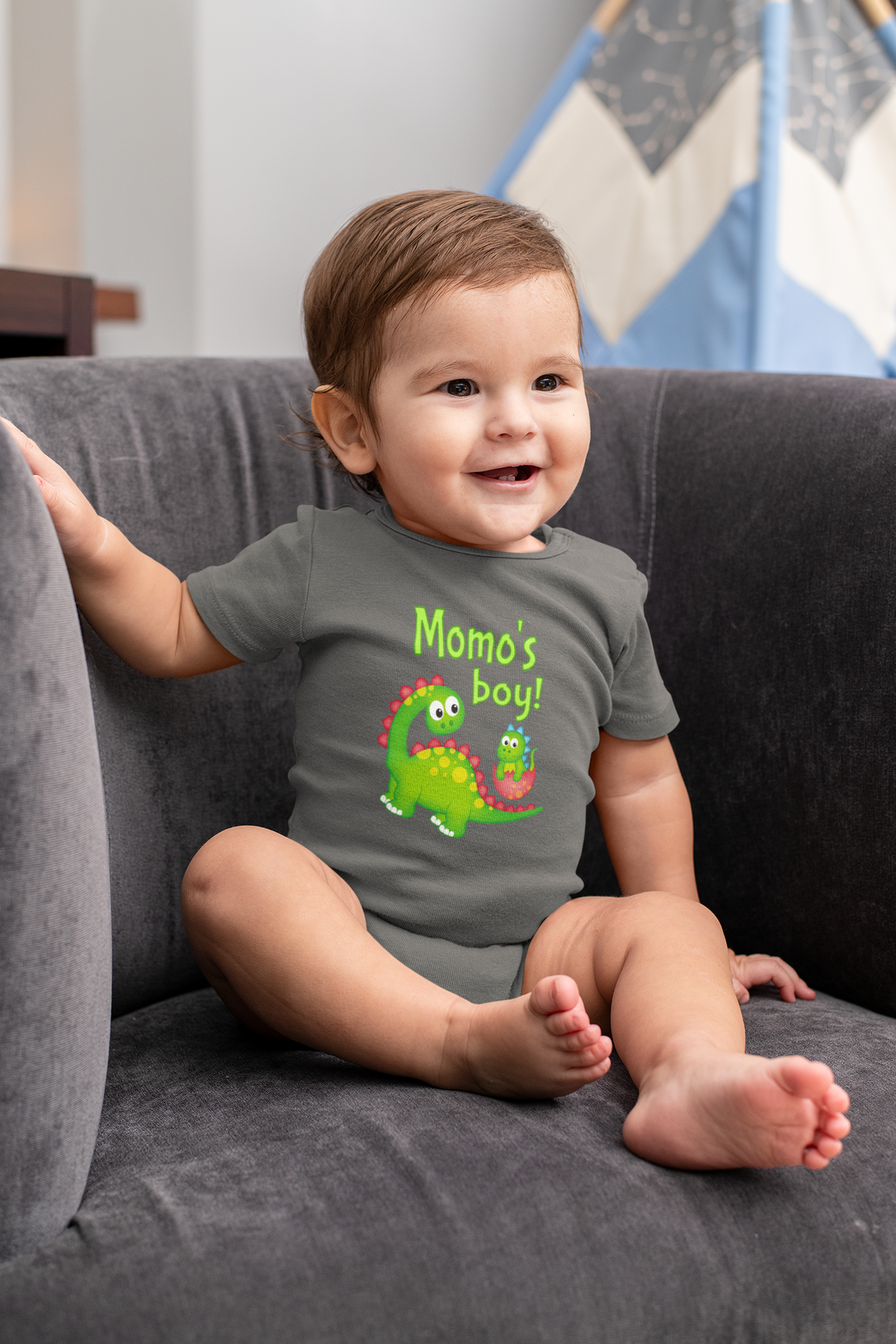 A smiling baby boy wearing a grey t-shirt with a dinosaur graphic and the text "Momo's boy!" sitting on a couch.