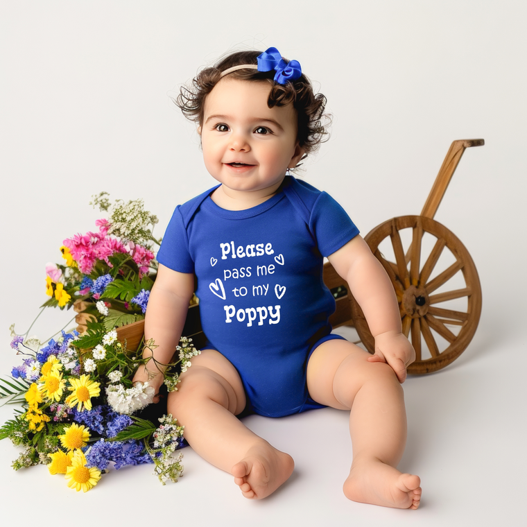A smiling baby girl wearing a blue onesie with the text "Please pass me to my Poppy" sits next to a wooden wagon filled with colorful flowers.