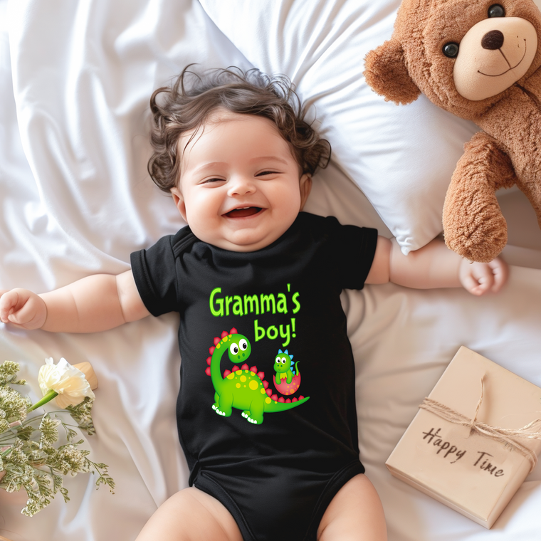 A smiling baby wearing a black t-shirt with a dinosaur graphic lies on a bed next to a stuffed teddy bear.