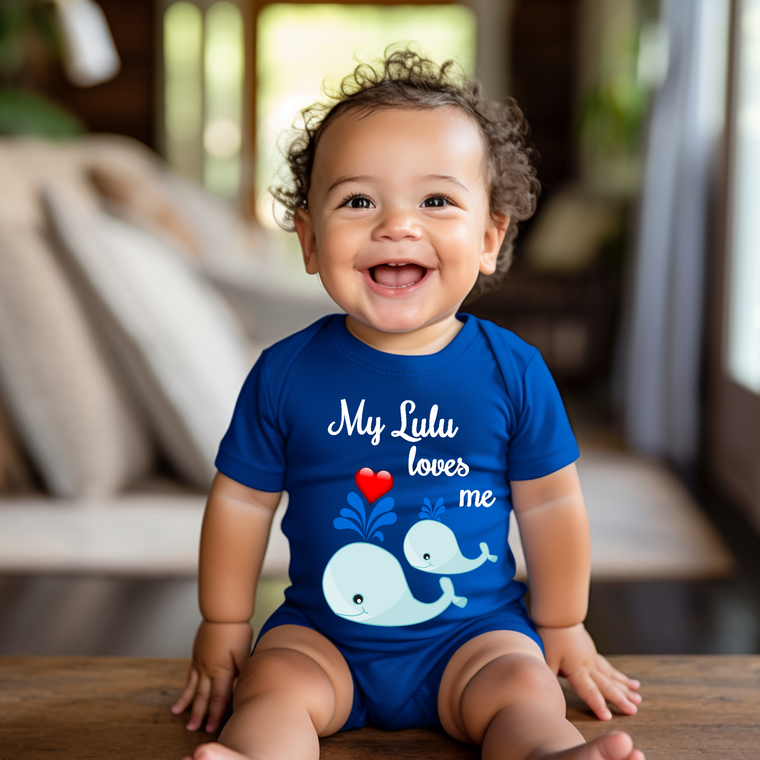 A smiling baby boy wearing a blue onesie with a whale design and the text "My Lulu loves me" is sitting on a wooden surface.
