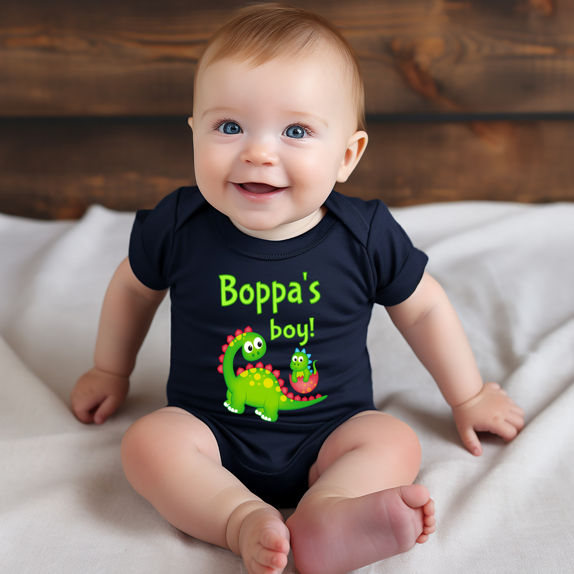 A smiling baby wearing a black shirt with a cartoon dinosaur and the text "Boppa's boy!"