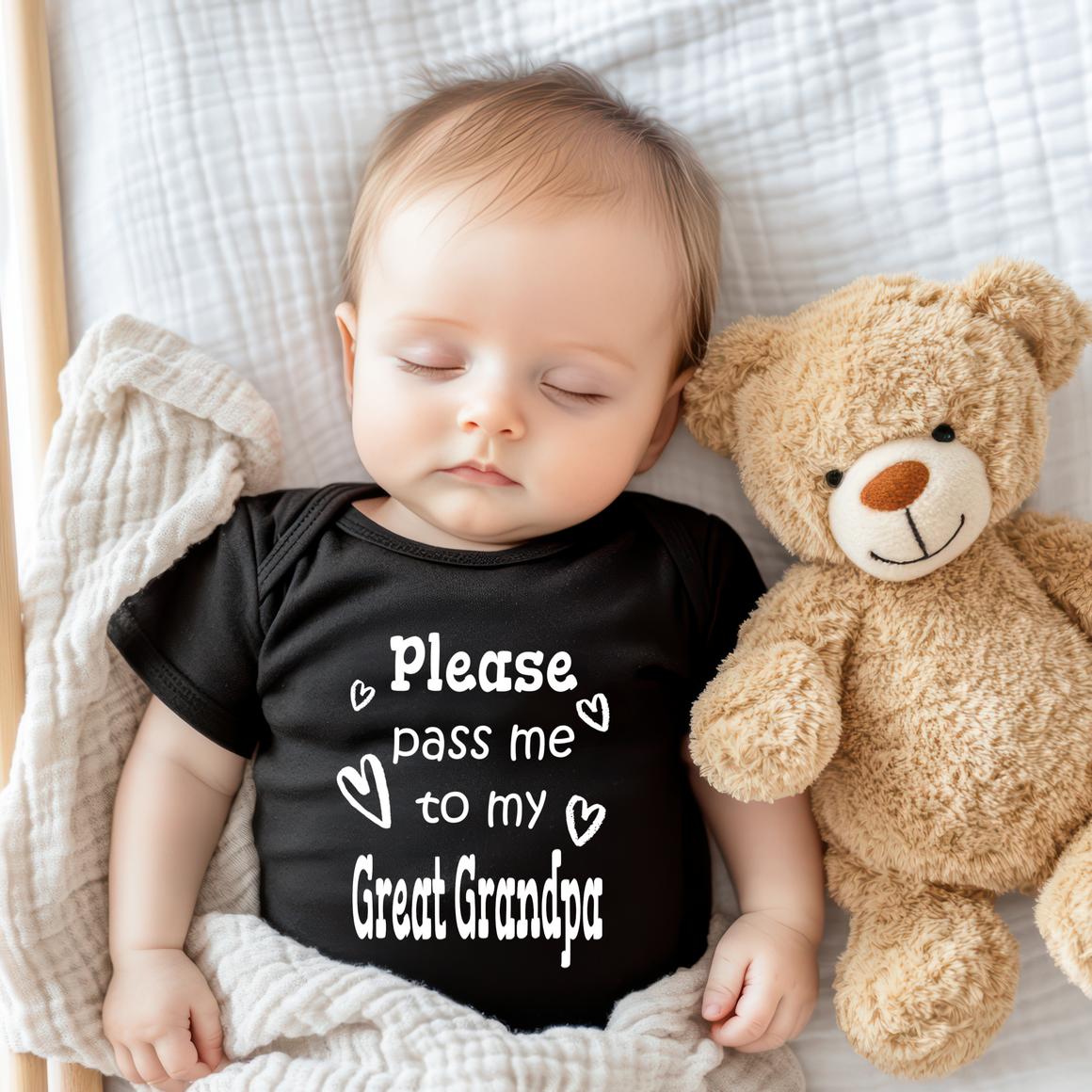 A sleeping baby wearing a black shirt with the text "Please pass me to my Great Grandpa" is lying next to a teddy bear on a bed.