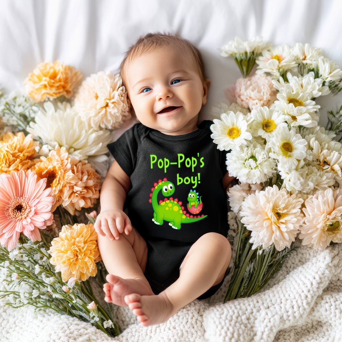 A smiling baby wearing a black shirt with a dinosaur graphic lies on a bed surrounded by colorful flowers.