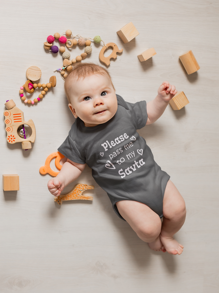 A baby lies on a white floor surrounded by various toys, including wooden blocks, a toy train, and a giraffe-shaped teething ring. The baby is wearing a gray onesie with the text "Please pass me to my Santa" printed on it.
