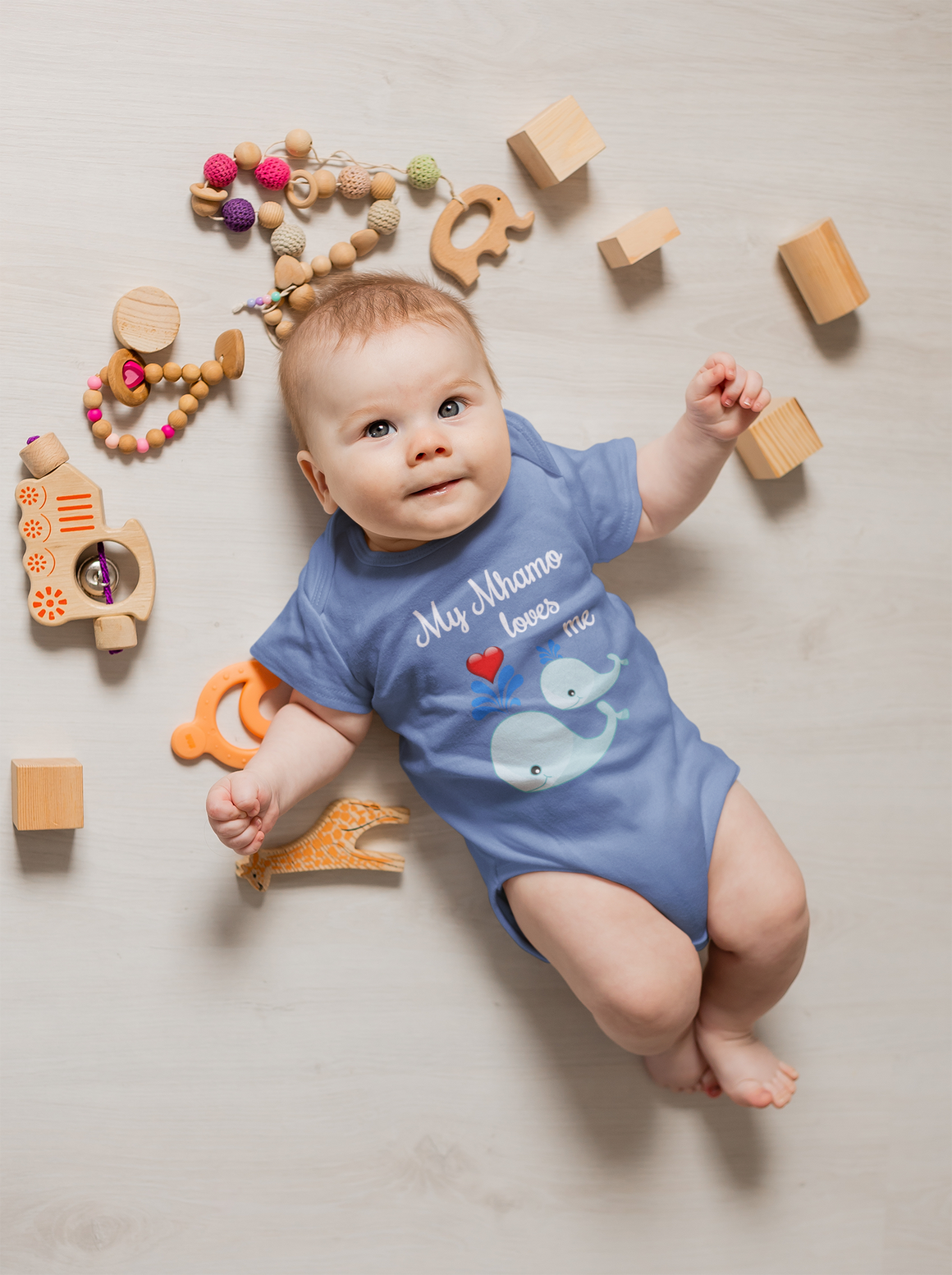 A baby lies on a wooden floor surrounded by various toys, including wooden blocks, a wooden train, and a wooden giraffe.