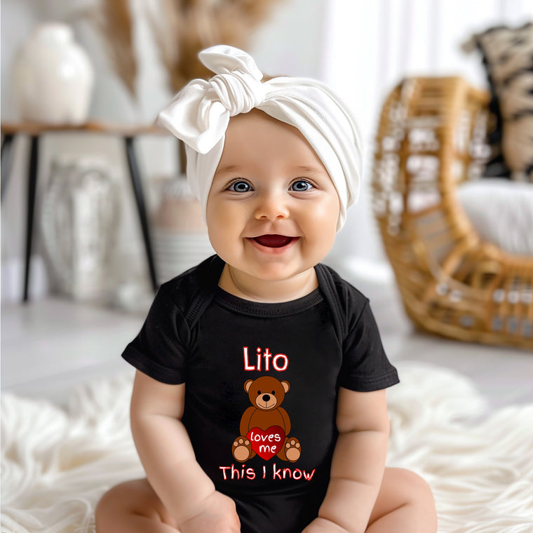 A smiling baby girl wearing a black t-shirt with a teddy bear graphic and the text "Lito loves me This I know" is sitting on a white blanket.