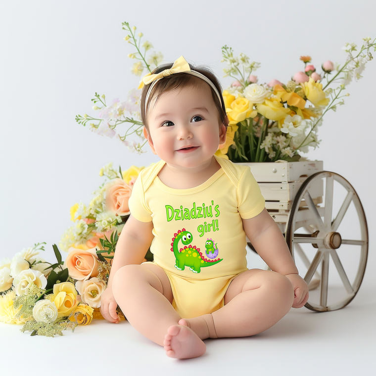 A smiling baby girl wearing a yellow onesie with a dinosaur on it, sitting on the floor in front of a wooden wagon filled with flowers.