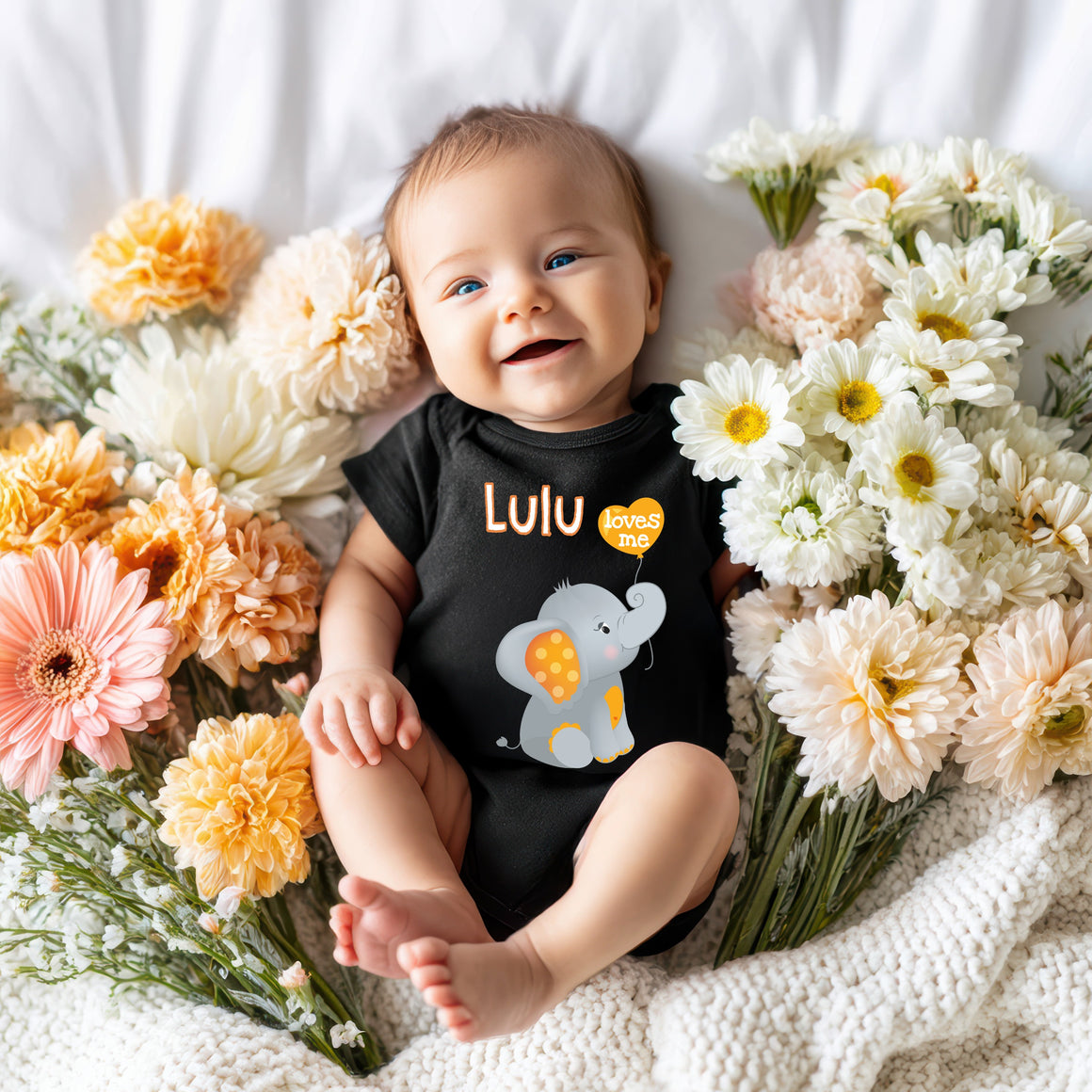 A smiling baby wearing a black shirt with an elephant design lies surrounded by a bouquet of colorful flowers.