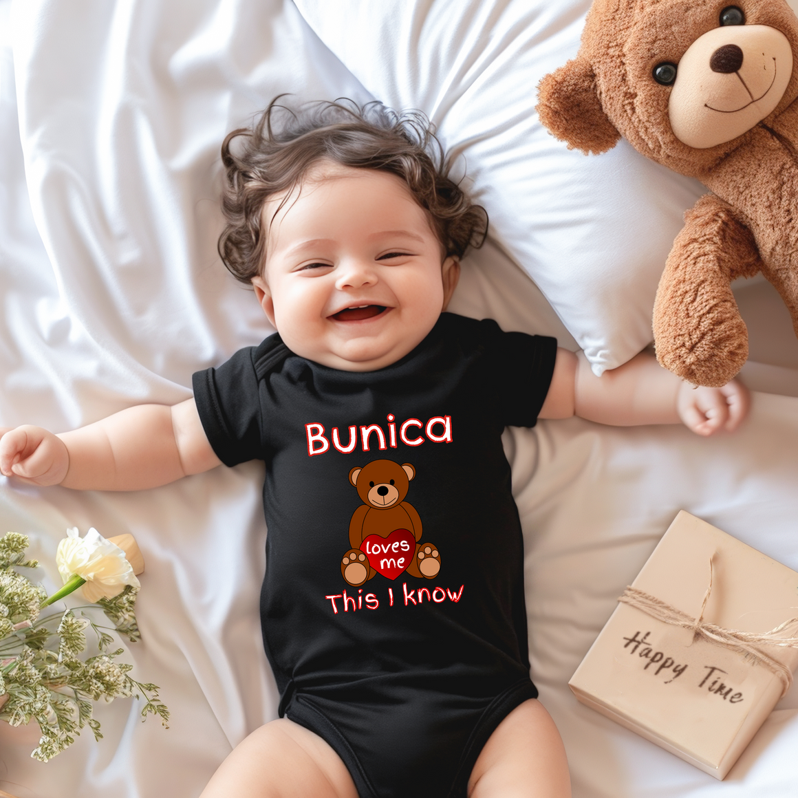 A smiling baby wearing a black onesie with a teddy bear and a book on a bed.