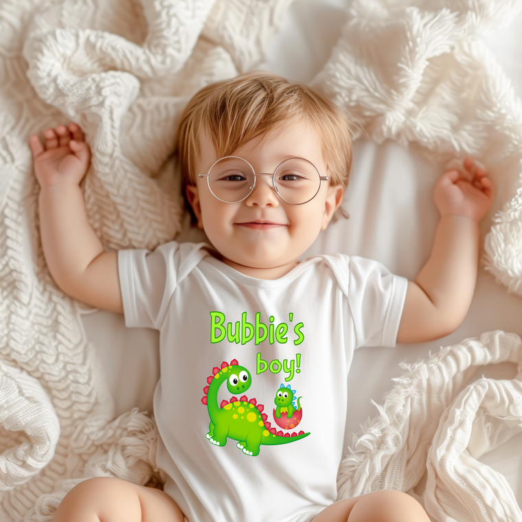 A smiling baby wearing a white onesie with a dinosaur graphic and the text "Bubbie's boy!" lying on a white blanket.