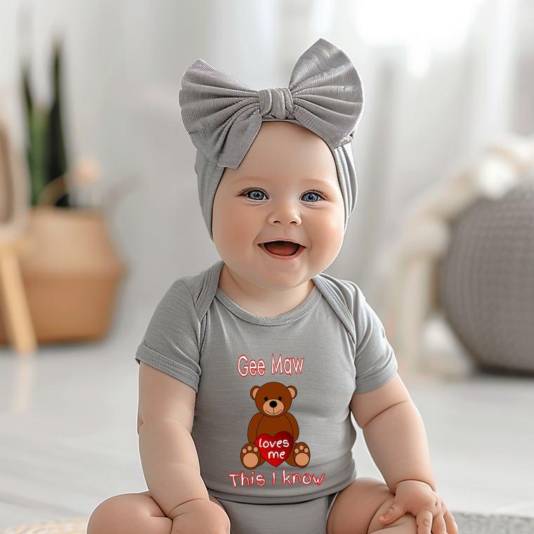 A smiling baby girl wearing a gray headband and a gray onesie with a teddy bear graphic and text.