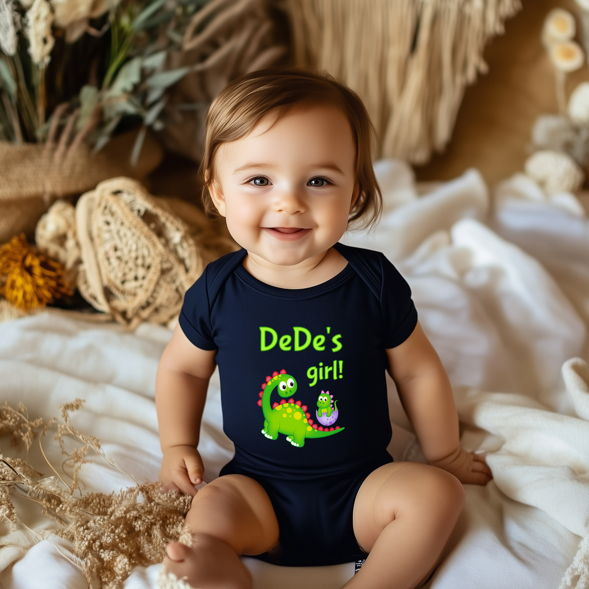 A smiling baby wearing a black t-shirt with a dinosaur graphic and the text "DeDe's girl!"