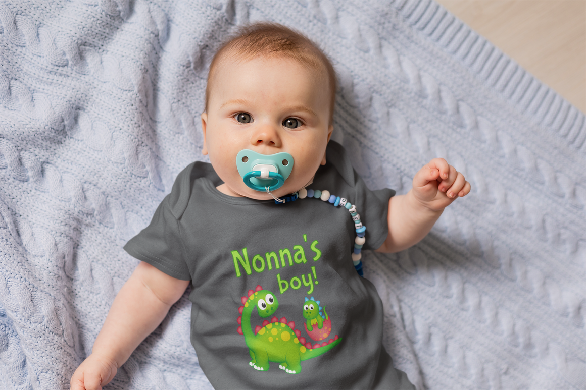 A baby wearing a gray t-shirt with a dinosaur design and the text "Nonna's boy!" lying on a blue blanket.