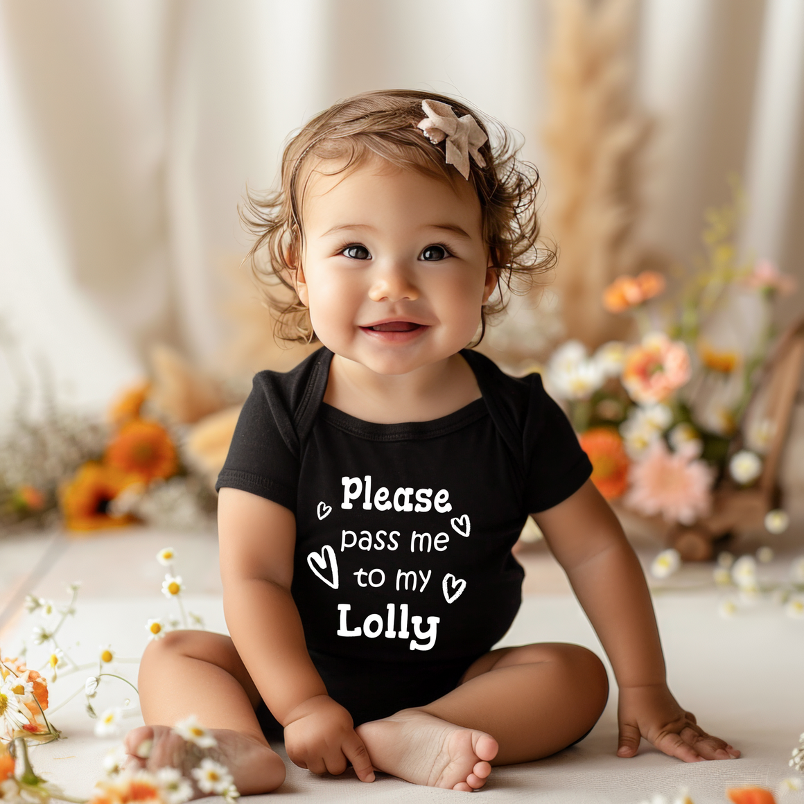 A smiling baby girl wearing a black onesie with the text "Please pass me to my Lolly" sits on the floor surrounded by flowers.