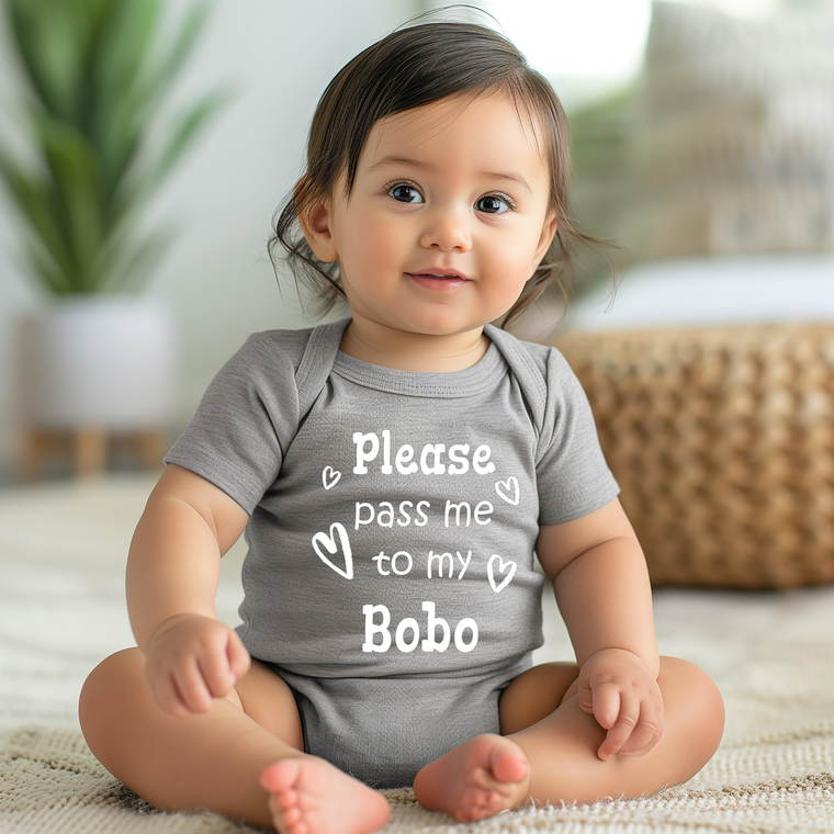 A smiling baby girl wearing a grey onesie with the text "Please pass me to my Bobo" printed on it.