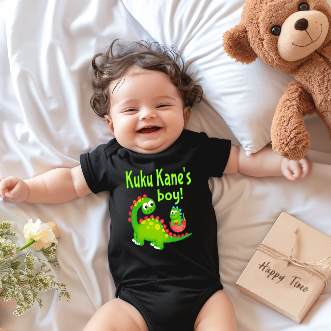 A smiling baby wearing a black onesie with a dinosaur design lies on a bed next to a stuffed teddy bear.