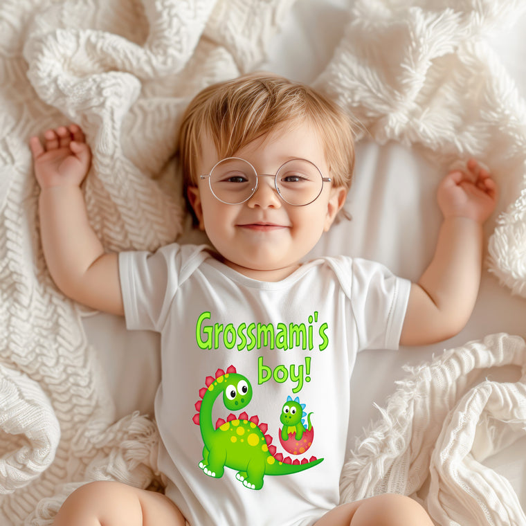 A smiling baby wearing a white onesie with a dinosaur graphic and the text "Grossmami's boy!" lying on a white blanket.