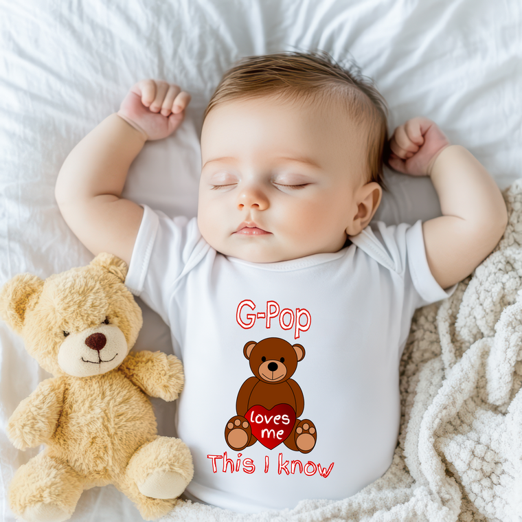A sleeping baby wearing a white onesie with a teddy bear and text printed on it.