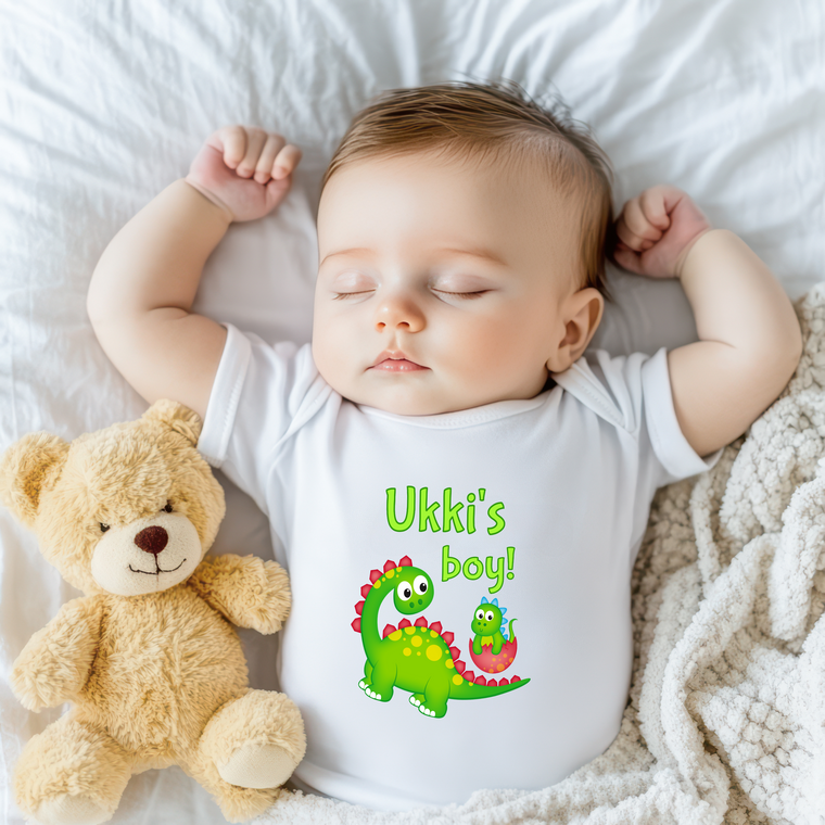 A sleeping baby wearing a white onesie with a dinosaur graphic and the text "Uki's boy!" next to a teddy bear.