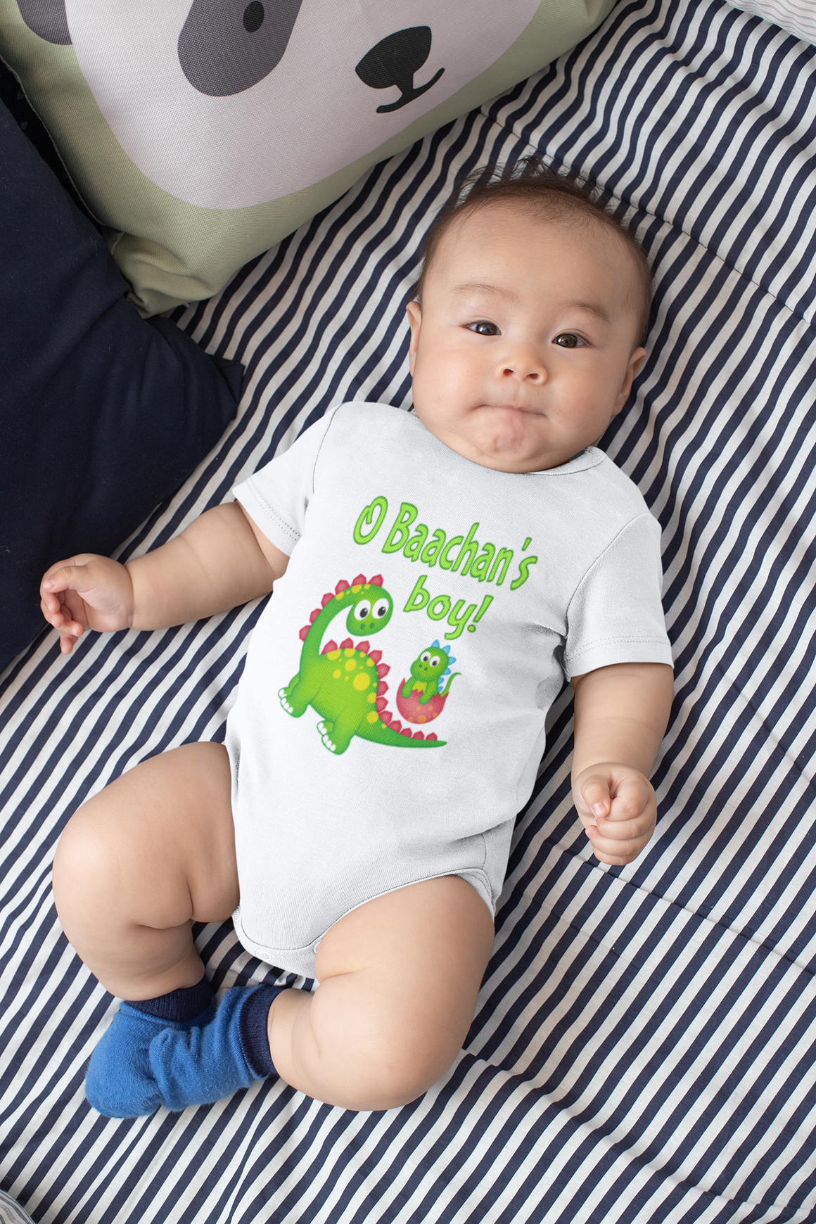 A baby wearing a white onesie with a dinosaur design and the text "O'Baker's Boy!" lying on a striped bed.