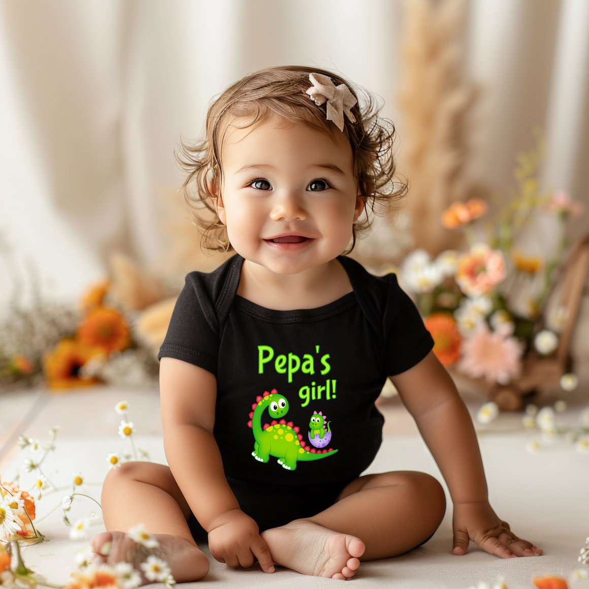 A smiling baby girl wearing a black shirt with a dinosaur and the text "Pepa's girl!" is sitting on the floor surrounded by flowers.