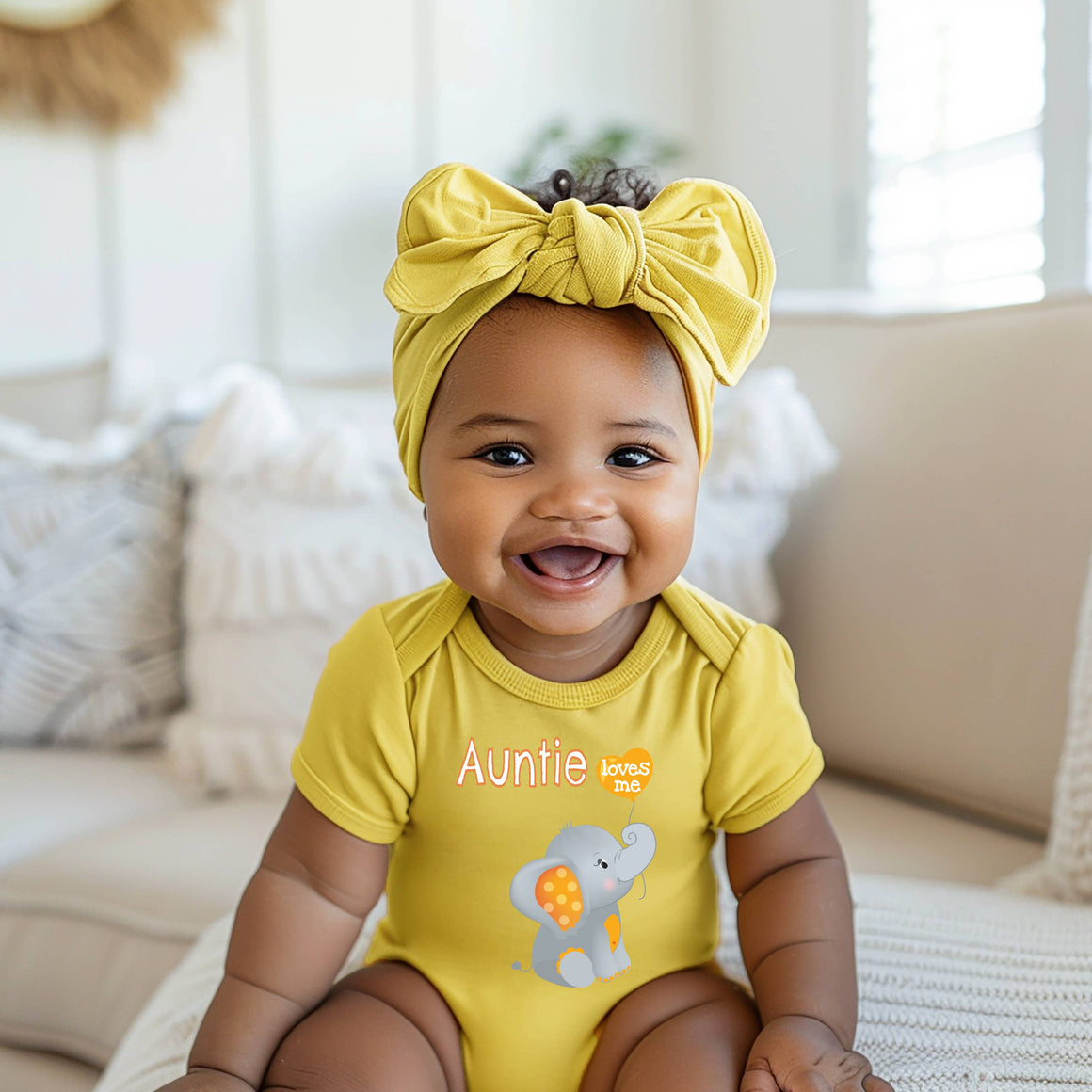 A smiling baby girl wearing a yellow outfit with a bow on her head and a yellow elephant graphic on her shirt, sitting on a couch.